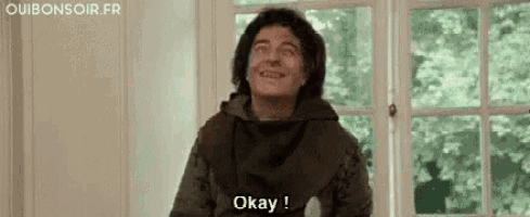 a man is sitting in front of a window and smiling while saying okay .
