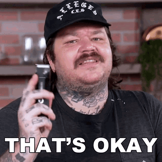 a man with tattoos is holding a hair clipper and says " that 's okay " on the bottom