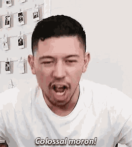 a man in a white shirt is making a funny face with his mouth open and says colossal moron .