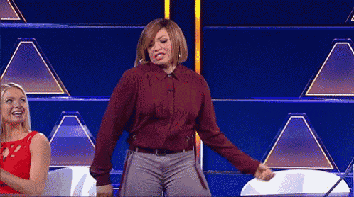 a woman in a maroon shirt is dancing on a television show while another woman looks on .