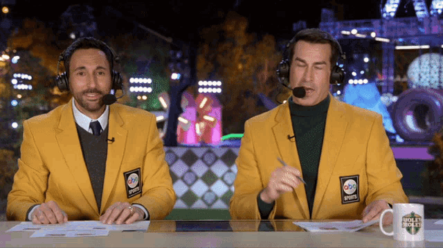 two men wearing yellow sports jackets sit at a table with a mug that says hockey world