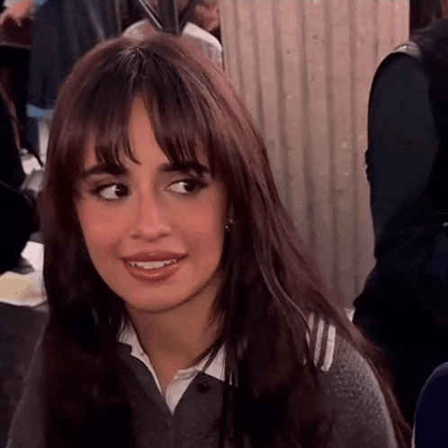 a woman with long brown hair and bangs is wearing a grey sweater and white shirt .
