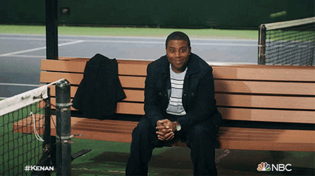 a man sits on a bench in front of a tennis court with the nbc logo visible