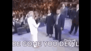 a man in a white suit is standing in front of a crowd and talking to a woman in a white dress .