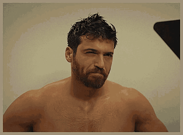 a shirtless man with a beard looks at the camera with his eyes closed