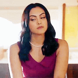 a woman in a purple dress and pearl necklace is sitting on a couch making a funny face .