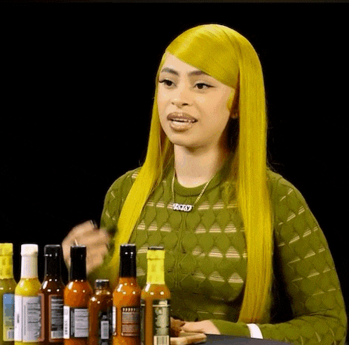 a woman with yellow hair is standing in front of a table full of hot sauce bottles .