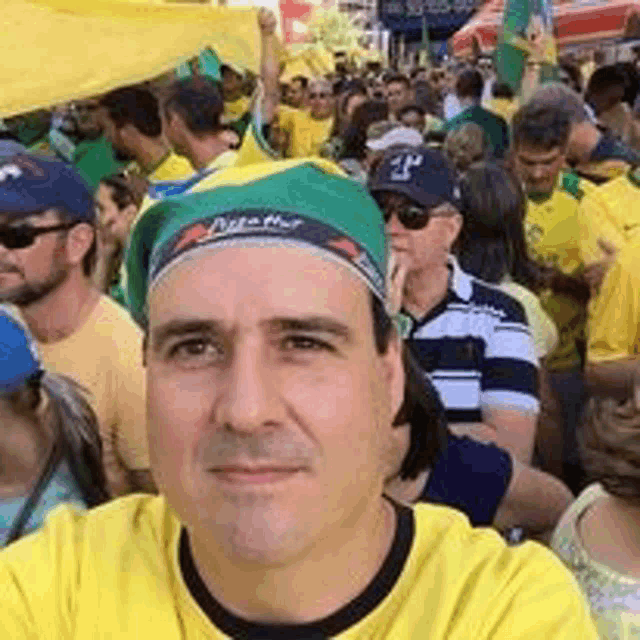 a man in a yellow shirt and green hat is taking a selfie in front of a crowd .