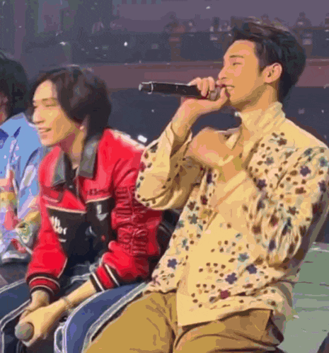 a man is singing into a microphone while sitting next to another man in a red jacket