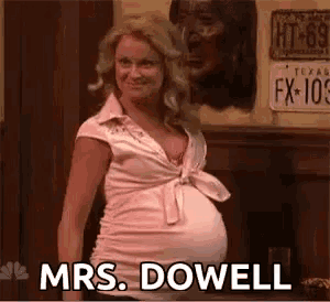 a pregnant woman in a pink shirt is standing in front of a wooden wall and says `` mrs. dowell '' .