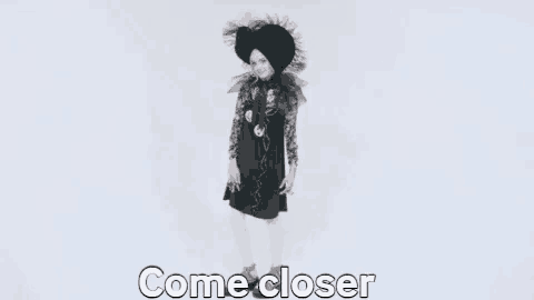 a woman in a black dress and white tights is standing in front of a white background and says `` come closer '' .