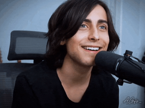 a young man with long hair is smiling in front of a microphone with a gif written on the bottom right