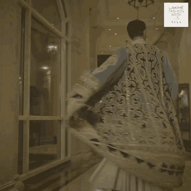 a man in a blue and gold dress is standing in a hallway with a lakme fashion week logo on the bottom