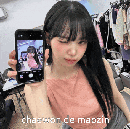 a woman in a pink tank top is holding a cell phone with a picture of herself on it and the caption chaewon de maozin