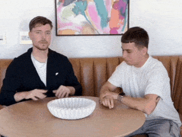 two men sit at a table with a bowl on it and a painting on the wall behind them