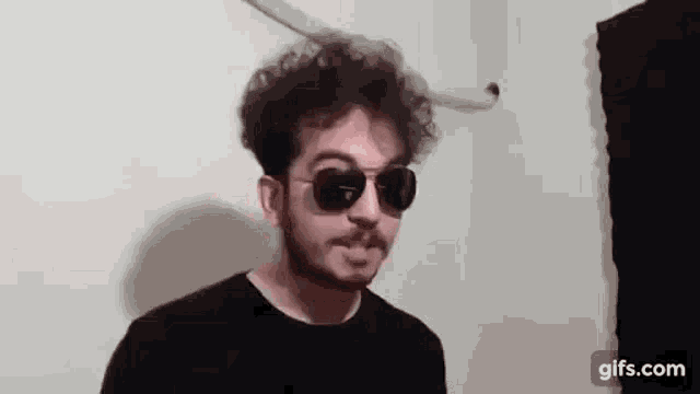 a man wearing sunglasses and a black shirt is standing in front of a wall .