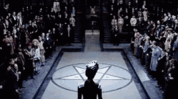 a woman in a crown stands in front of a pentagram in a circle surrounded by people