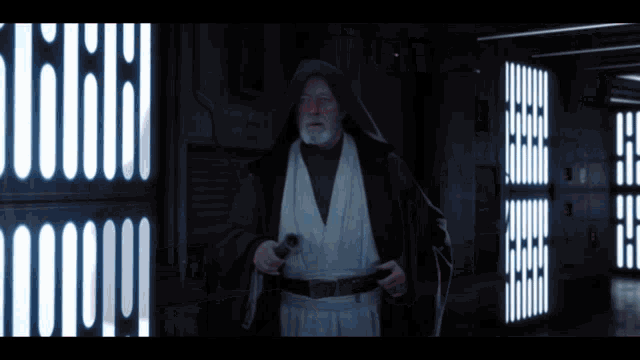 a man in a hooded robe holds a lightsaber in a dark hallway