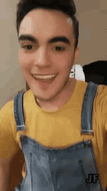 a young man wearing overalls and a yellow shirt is smiling and taking a selfie .