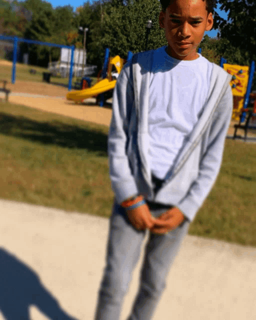 a young boy in a white shirt and grey pants stands in front of a playground