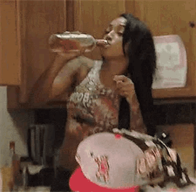 a woman is drinking from a bottle in a kitchen while wearing a baseball cap .