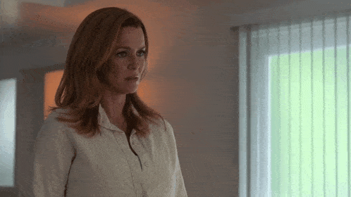a woman with red hair is standing in front of a window with vertical blinds .