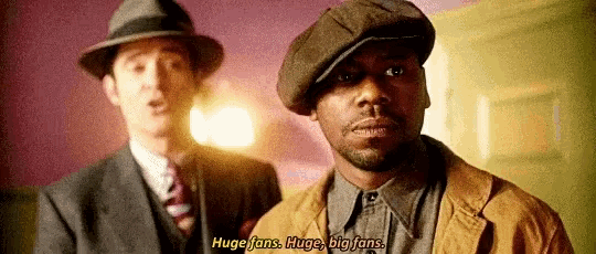 two men in suits and hats are standing next to each other and one of them is talking about huge fans .