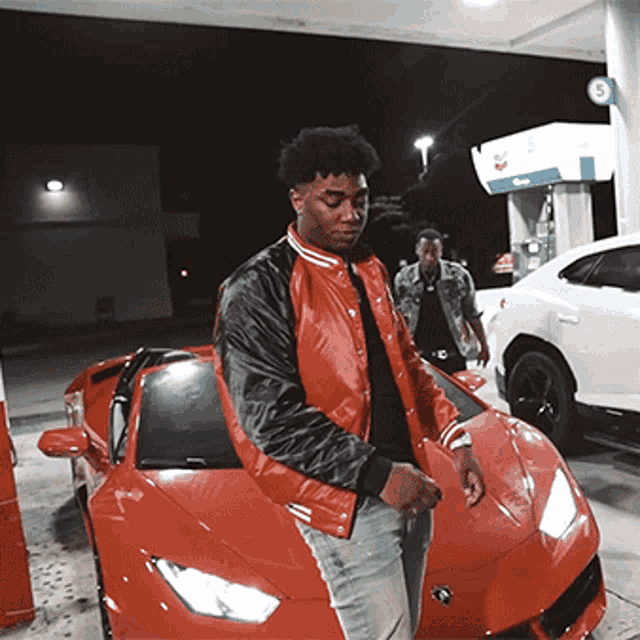 a man in a red jacket is standing next to a red car at a gas station