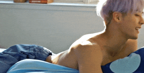 a shirtless man with purple hair is laying on a bed with a blue pillow
