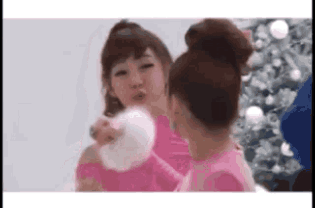two girls are standing next to each other in front of a christmas tree . one of the girls is wearing a pink shirt .