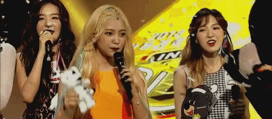 three girls are standing next to each other on a stage holding microphones and a stuffed animal .