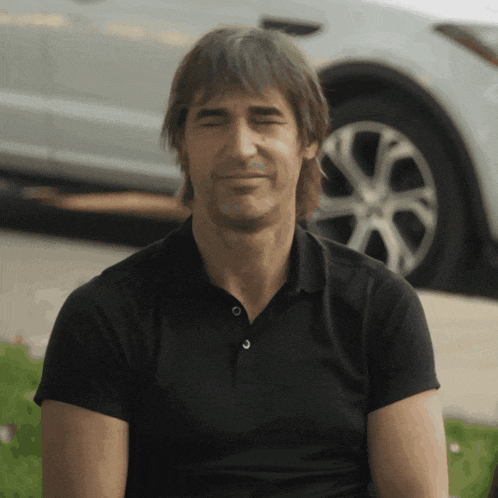 a man wearing a black polo shirt is smiling in front of a silver car