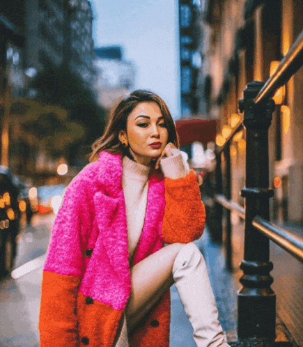a woman wearing a bright pink and orange coat sits on a sidewalk