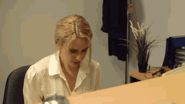 a woman in a white shirt sits at a desk with a plant in the background