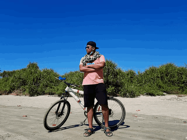 a man in a pink shirt stands next to a bicycle on the beach