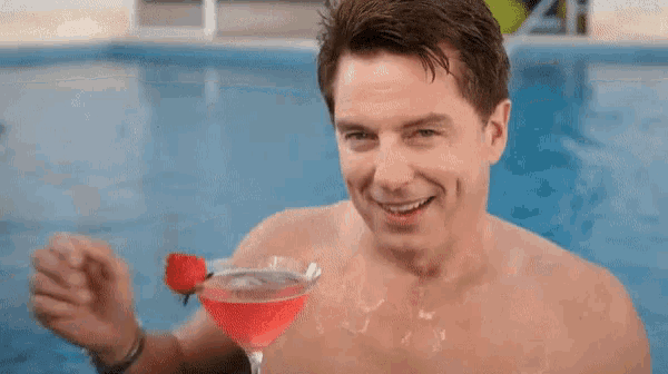 a shirtless man in a swimming pool holds a red cocktail