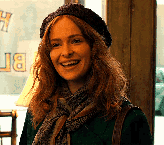 a woman wearing a hat and scarf smiles in front of a window that says jb on it