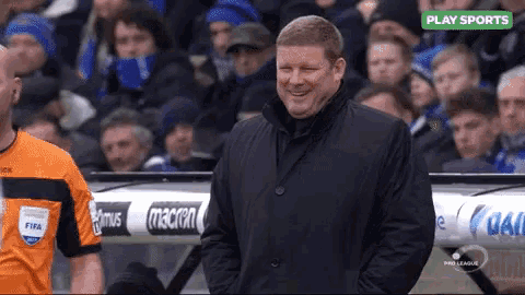 a man in a black jacket is smiling in front of a crowd and a sign that says play sports