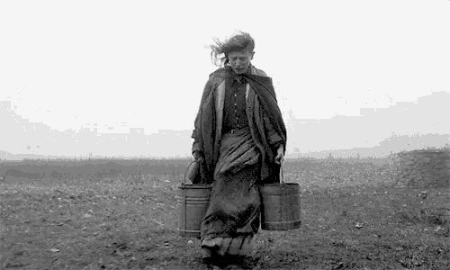 a black and white photo of a woman walking in a field carrying two buckets .