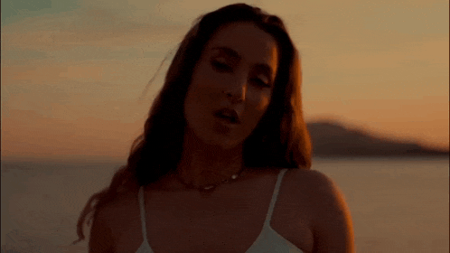 a woman in a white tank top and necklace is standing on the beach at sunset .
