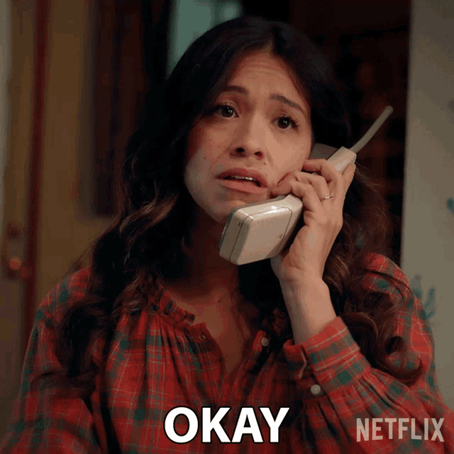 a woman in a plaid shirt is talking on a phone and the word okay is on the bottom