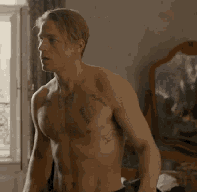 a shirtless man with tattoos on his chest is standing in a room in front of a mirror .