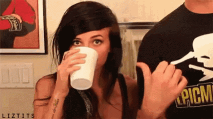 a woman is drinking from a styrofoam cup while a man in a skull shirt looks on .