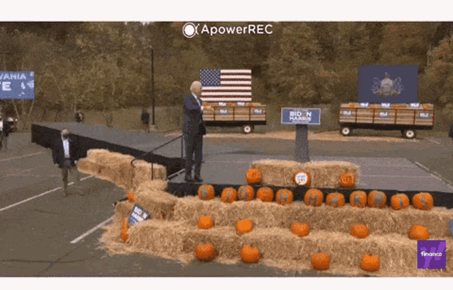 a man stands on a stage surrounded by pumpkins and hay bales with a biden sign behind him