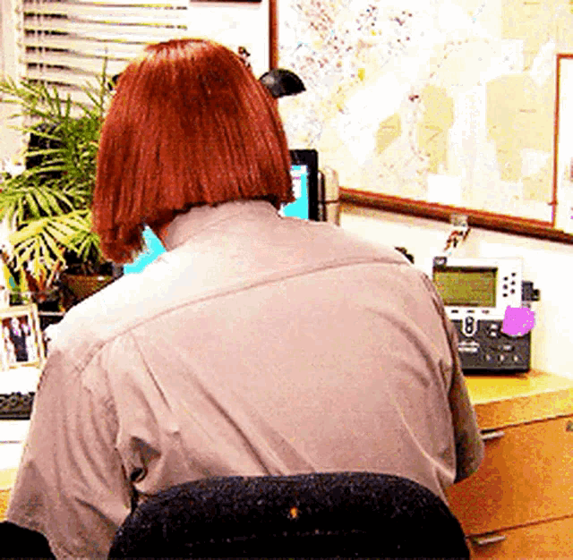 a woman with red hair is sitting at a desk with a phone and a computer .