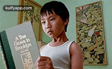 a young boy in a white tank top is reading a book in a room .