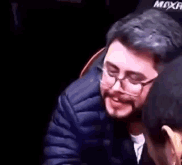 a man with glasses and a beard is sitting next to another man in a dark room .