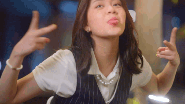 a woman wearing a vest and a pearl necklace sticks her tongue out