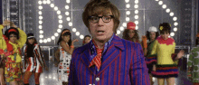 a man in a purple suit and tie is standing in front of a group of women in colorful costumes .