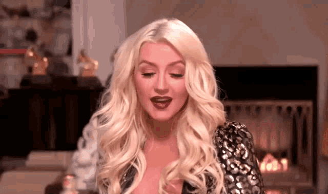 a woman with blonde hair and purple lipstick is standing in front of a fireplace in a room .